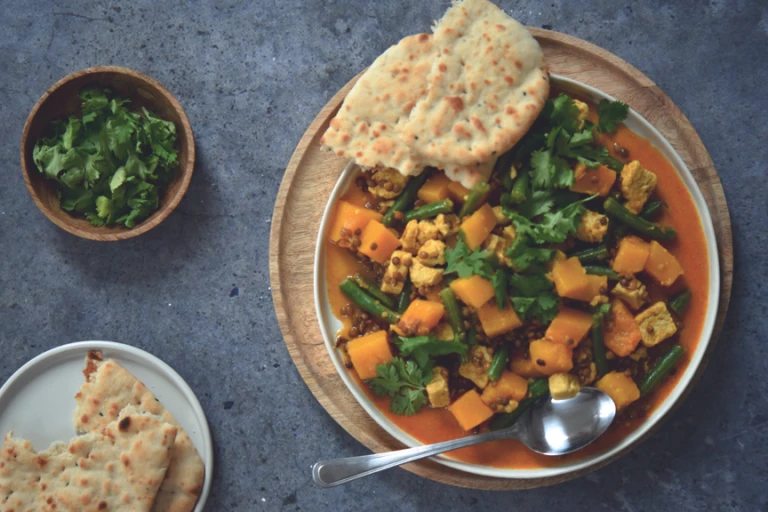 A plate of yellow curry with Quorn pieces, lentils and squash sat beside a bowl of coriander & a plate of naan bread.