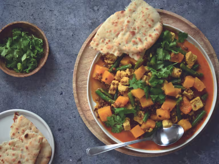 A plate of yellow curry with Quorn pieces, lentils and squash sat beside a bowl of coriander & a plate of naan bread.