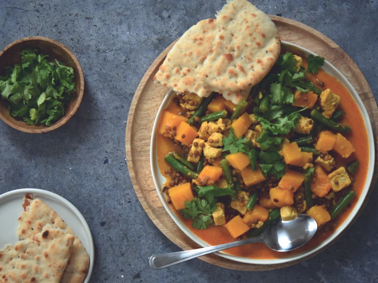 A plate of yellow curry with Quorn pieces, lentils and squash sat beside a bowl of coriander & a plate of naan bread.