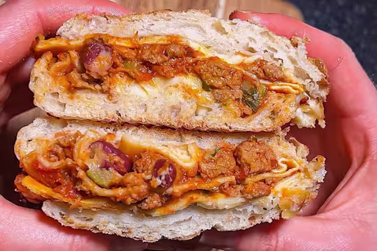 Someone holding a sandwich cut open filled with Quorn Vegetarian Mince Lasagne over a wooden board.