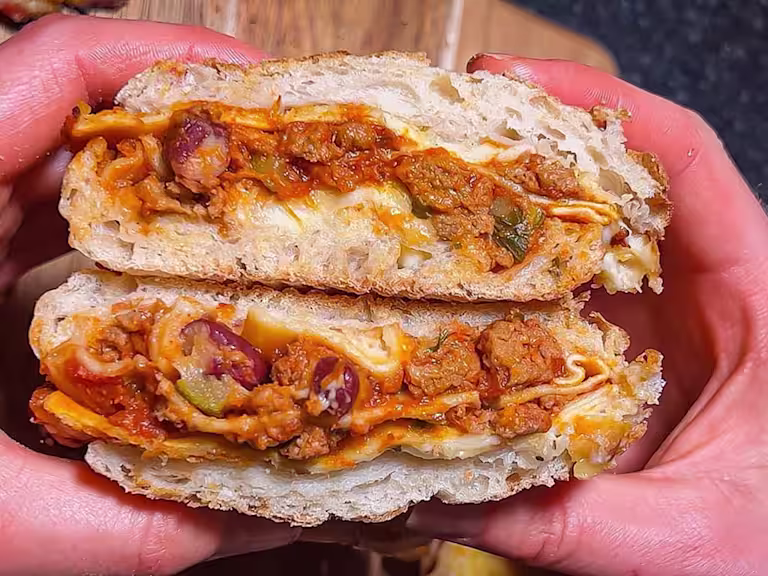 Someone holding a sandwich cut open filled with Quorn Vegetarian Mince Lasagne over a wooden board.