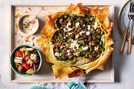 Feta and spinach Spanakopita on a wooden serving board with a yoghurt dip and side dish of vegetables.