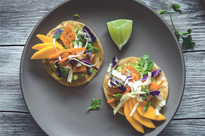 Two tostadas topped with Quorn Fishless Sticks, avocado, mango, and salw with a wedge of lime on the side.