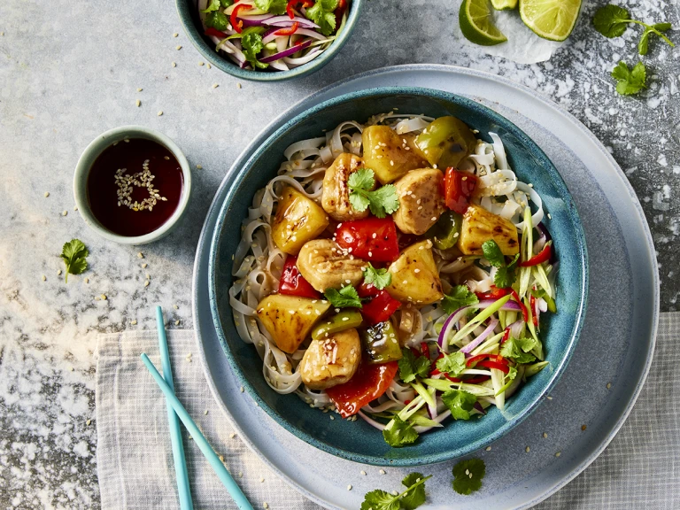 A bowl of Quorn vegetarian sweet and sour chicken with sesame noodles with Quorn Fillet Pieces visible on top with a side of sauce. 