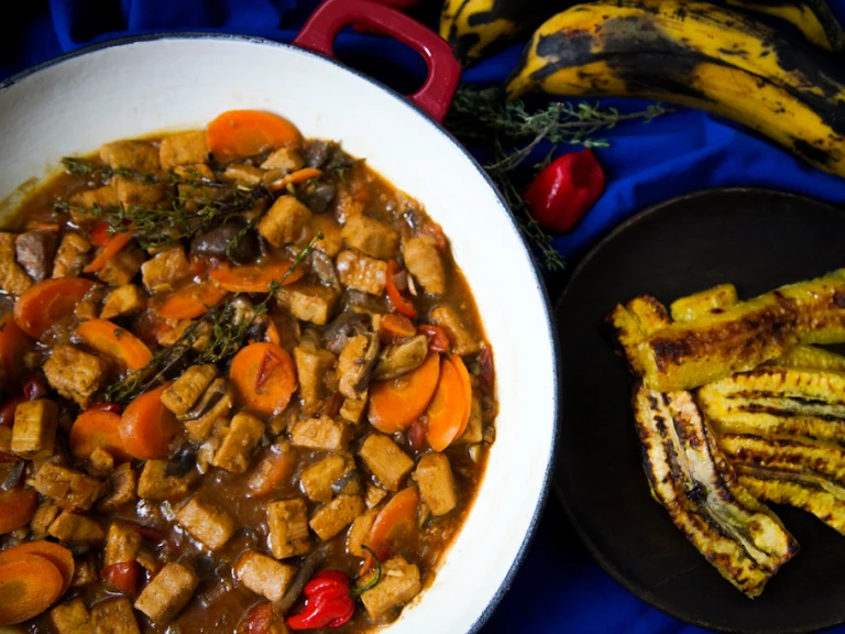 A red Dutch oven filled with Jamaican Brown Stew made with Quorn Vegan Pieces, sliced carrots, scotch bonnet chilli, and thyme on a blue background with a dish of plantain on the side.