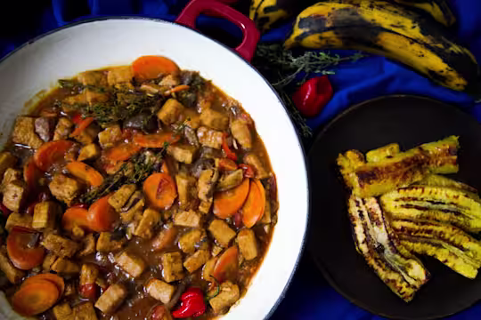 A red Dutch oven filled with Jamaican Brown Stew made with Quorn Vegan Pieces, sliced carrots, scotch bonnet chilli, and thyme on a blue background with a dish of plantain on the side.