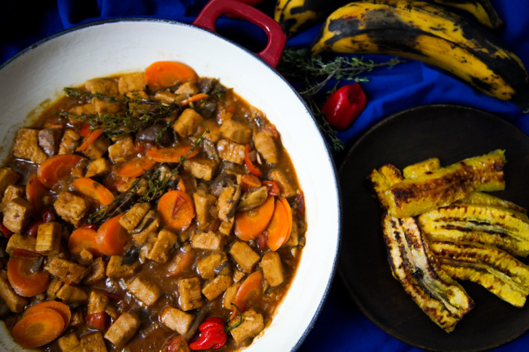 A red Dutch oven filled with Jamaican Brown Stew made with Quorn Vegan Pieces, sliced carrots, scotch bonnet chilli, and thyme on a blue background with a dish of plantain on the side.