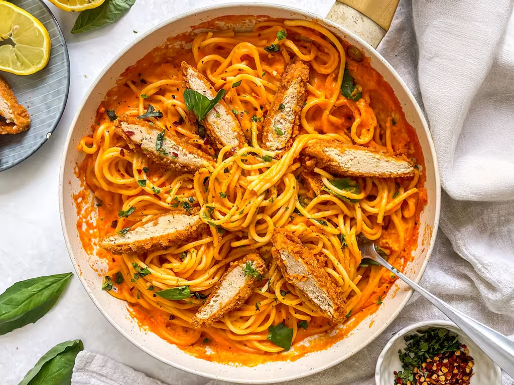 Bowl filled with red pepper spaghetti and topped with sliced Quorn Crunchy Fillet Burgers and fresh basil. The pack and garnish such as lemon and chilli flakes are also in the background. 