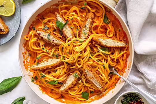 Bowl filled with red pepper spaghetti and topped with sliced Quorn Crunchy Fillet Burgers and fresh basil. The pack and garnish such as lemon and chilli flakes are also in the background.