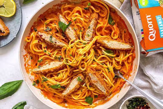 Bowl filled with red pepper spaghetti and topped with sliced Quorn Spicy Buffalo Fillets and fresh basil. The pack and garnish such as lemon and chilli flakes are also in the background.