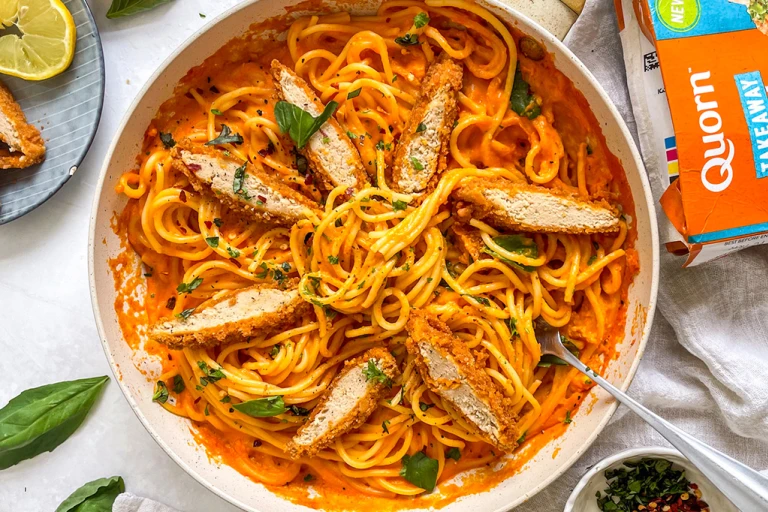 Bowl filled with red pepper spaghetti and topped with sliced Quorn Spicy Buffalo Fillets and fresh basil. The pack and garnish such as lemon and chilli flakes are also in the background. 