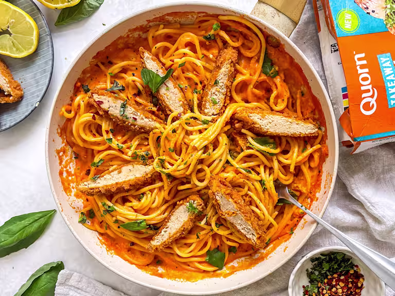Bowl filled with red pepper spaghetti and topped with sliced Quorn Spicy Buffalo Fillets and fresh basil. The pack and garnish such as lemon and chilli flakes are also in the background.