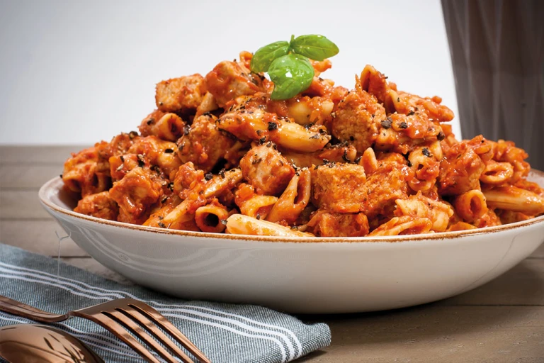 A bowl of one pot pasta made with penne pasta and Quorn Vegan Pieces in a tomato-based sauce garnished with a sprig of basil.