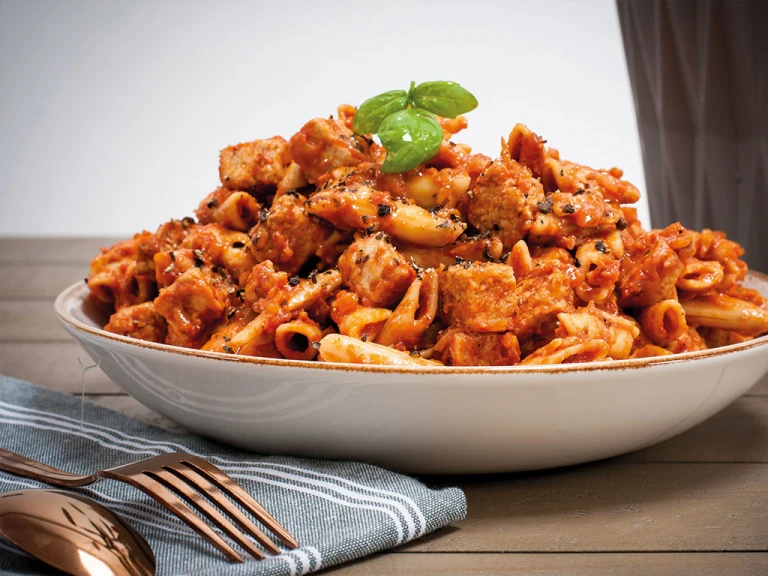 A bowl of one pot pasta made with penne pasta and Quorn Vegan Pieces in a tomato-based sauce garnished with a sprig of basil.