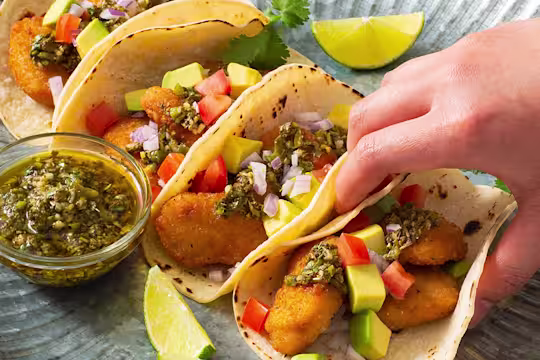 Four tacos filled with air-fried Quorn Crispy Nuggets and diced avocado, tomato, and onion, and topped with chimichurri sauce with extra sauce and lime wedges on the side.