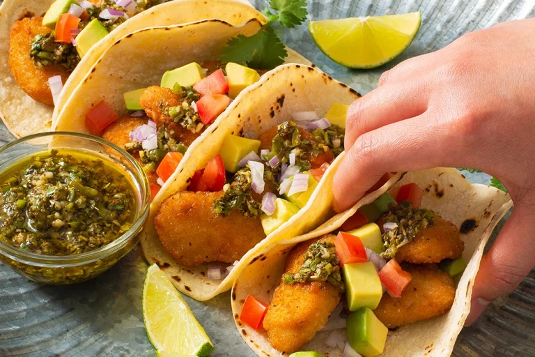 Four tacos filled with air-fried Quorn Crispy Nuggets and diced avocado, tomato, and onion, and topped with chimichurri sauce with extra sauce and lime wedges on the side.