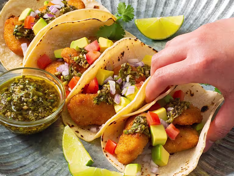 Four tacos filled with air-fried Quorn Crispy Nuggets and diced avocado, tomato, and onion, and topped with chimichurri sauce with extra sauce and lime wedges on the side.