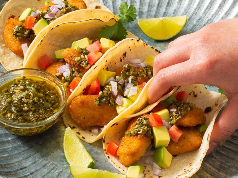 Four tacos filled with air-fried Quorn Crispy Nuggets and diced avocado, tomato, and onion, and topped with chimichurri sauce with extra sauce and lime wedges on the side.