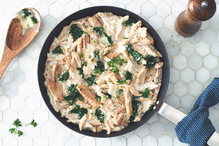 A pan full of penne in alfredo sauce with kale, Quorn Strips, and Parmesan cheese on a white tile background with a wooden spoon and pepper grinder on either side.