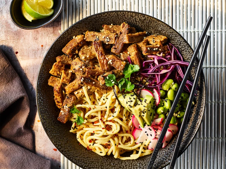 Quorn steak strips with sesame noodles served in a black bowl with black chopsticks and sliced lime on the side.