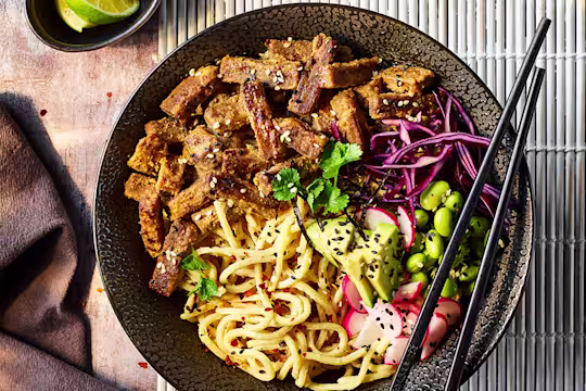 Quorn steak strips with sesame noodles served in a black bowl with black chopsticks and sliced lime on the side.