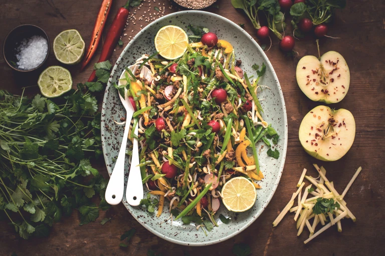 A serving plate of Easy Thai Salad with Quorn Mince sat on a table containing an apple, salt, chilli, radishes and herbs.