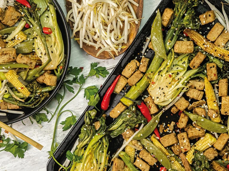 A traybake made with Quorn Pieces, bok choy, baby corn, sugar snap peas, chillies, bean sprouts, and tenderstem broccoli topped with sesame seeds shown plated and on the roasting tray.