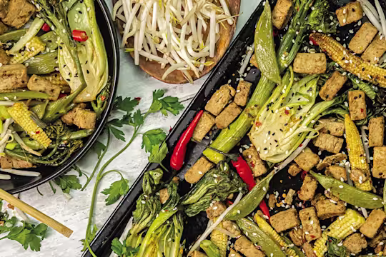 A traybake made with Quorn Pieces, bok choy, baby corn, sugar snap peas, chillies, bean sprouts, and tenderstem broccoli topped with sesame seeds shown plated and on the roasting tray.
