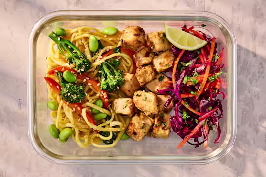 A glass meal prep container holding Vegetarian Satay Noodles with Quorn Pieces and a fresh cabbage slaw.
