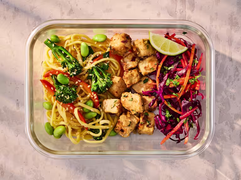 A glass meal prep container holding Vegetarian Satay Noodles with Quorn Pieces and a fresh cabbage slaw.