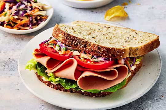 Quorn vegetarian ham slices in a sandwich with tomato and homemade coleslaw on seeded bread on a white plate.