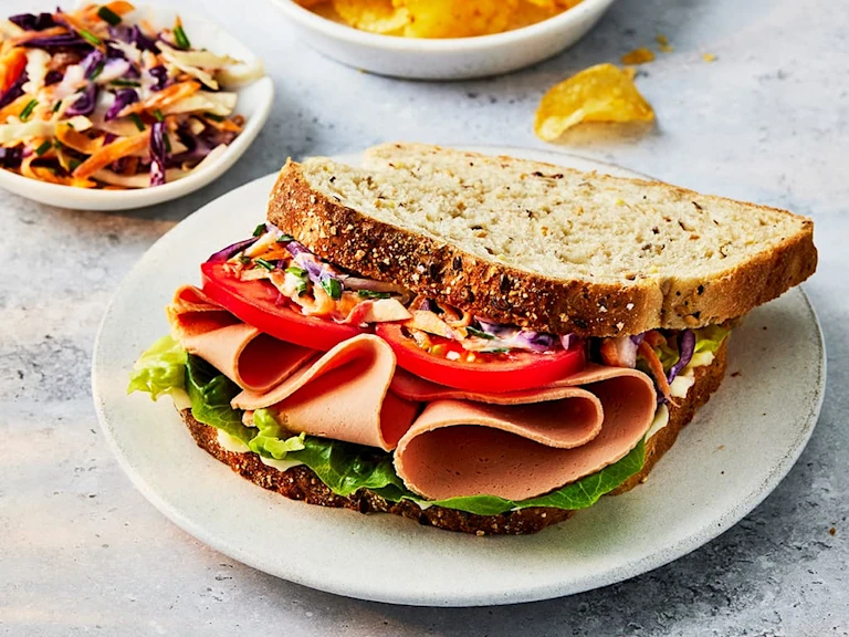 Quorn vegetarian ham slices in a sandwich with tomato and homemade coleslaw on seeded bread on a white plate.