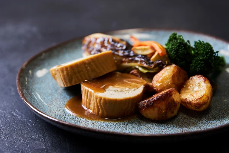 Two slices of a Quorn Roast stuffed with vegetarian stuffing served with roasted potatoes, roasted carrots and parsnips, broccoli, and roasted cabbage on the side and topped with gravy.
