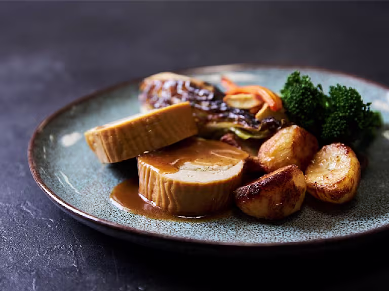 Two slices of a Quorn Roast stuffed with vegetarian stuffing served with roasted potatoes, roasted carrots and parsnips, broccoli, and roasted cabbage on the side and topped with gravy.