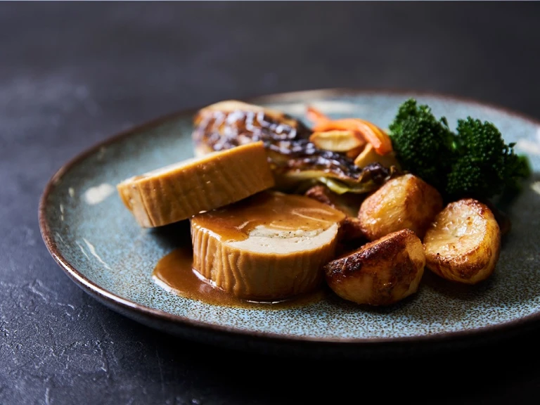 Two slices of a Quorn Roast stuffed with vegetarian stuffing served with roasted potatoes, roasted carrots and parsnips, broccoli, and roasted cabbage on the side and topped with gravy.