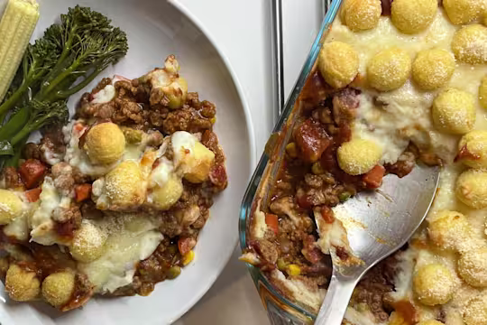 Vegetarian Shepherd’s Pie topped with potato pops in a dish with a serving spoon, plated with tenderstem broccoli and baby corn.
