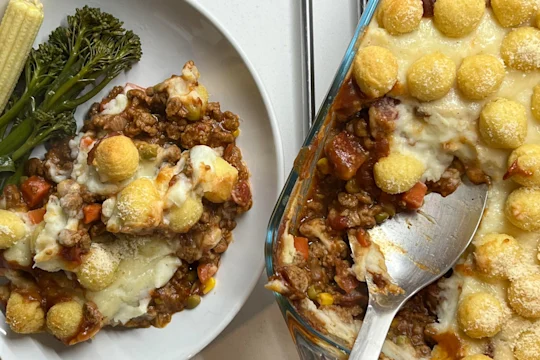 Vegetarian Shepherd’s Pie topped with potato pops in a dish with a serving spoon, plated with tenderstem broccoli and baby corn.