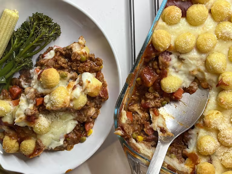 Vegetarian Shepherd’s Pie topped with potato pops in a dish with a serving spoon, plated with tenderstem broccoli and baby corn.