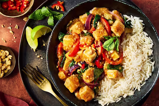 Quorn Pieces in a Massaman curry sauce with chopped peppers and onion served with white rice in a black bowl with lime garnish.