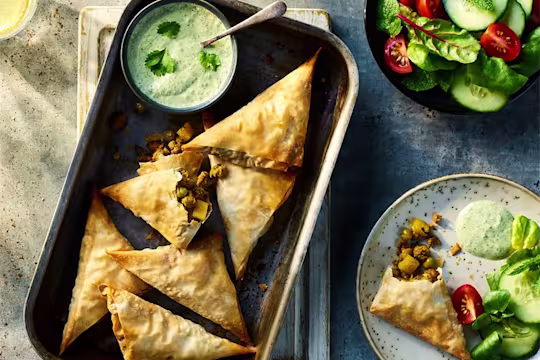 Six Vegetarian Samosas made using Quorn Vegetarian Mince served on a baking tray with an additional veggie samosa served on a plate to the side. They are served alongside mint and coriander raita.