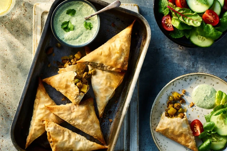 Six Vegetarian Samosas made using Quorn Vegetarian Mince served on a baking tray with an additional veggie samosa served on a plate to the side. They are served alongside mint and coriander raita. 