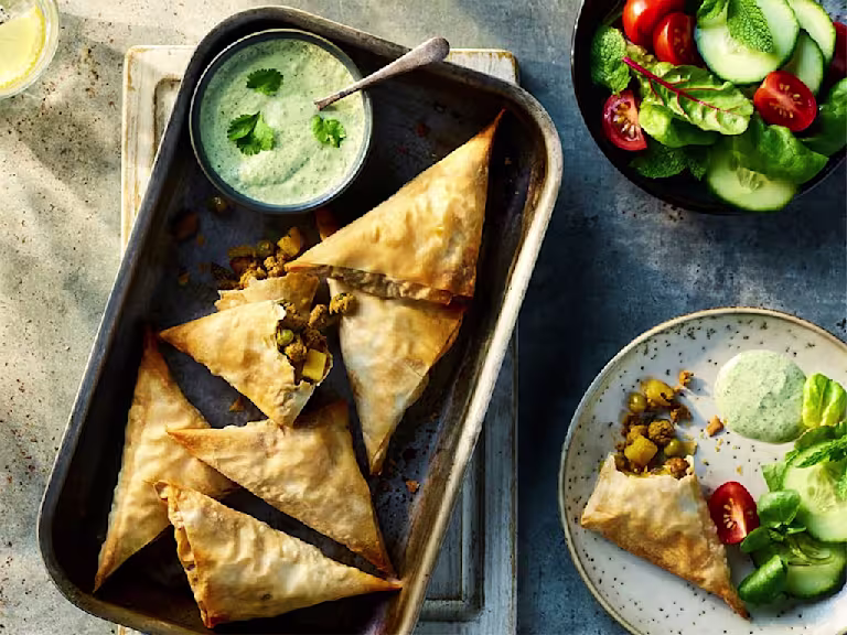 Six Vegetarian Samosas made using Quorn Vegetarian Mince served on a baking tray with an additional veggie samosa served on a plate to the side. They are served alongside mint and coriander raita.