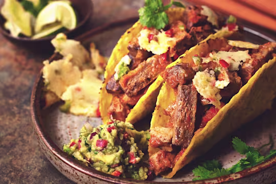 Two hard shell tacos filled with Quorn Vegetarian Steak Strips and queso fundido garnished with coriander and served on a black plate with guacamole on the side.