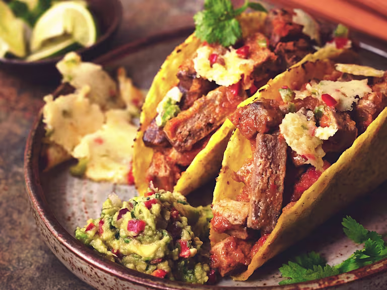 Two hard shell tacos filled with Quorn Vegetarian Steak Strips and queso fundido garnished with coriander and served on a black plate with guacamole on the side.
