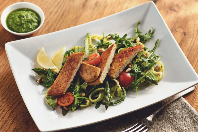 A sliced Quorn Fillet atop a bed of pasta with asparagus, arugula, and cherry tomatoes with two wedges of lemon and pesto on the side.