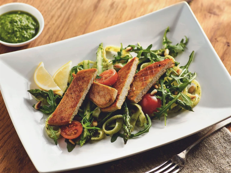 A sliced Quorn Fillet atop a bed of pasta with asparagus, arugula, and cherry tomatoes with two wedges of lemon and pesto on the side.