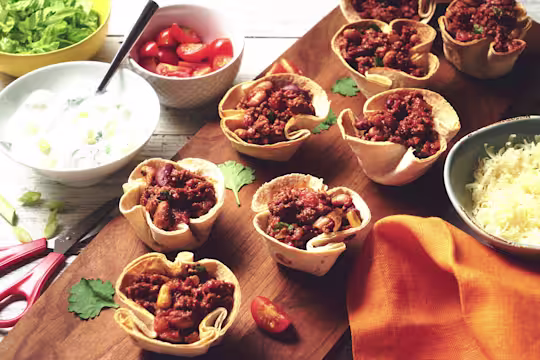 Vegetarian Quorn Mince Chilli filled in taco cups served on a wooden board with a side of cheese, yoghurt and cherry tomatoes