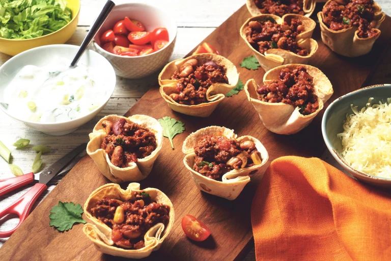 Vegetarian Quorn Mince Chilli filled in taco cups served on a wooden board with a side of cheese, yoghurt and cherry tomatoes