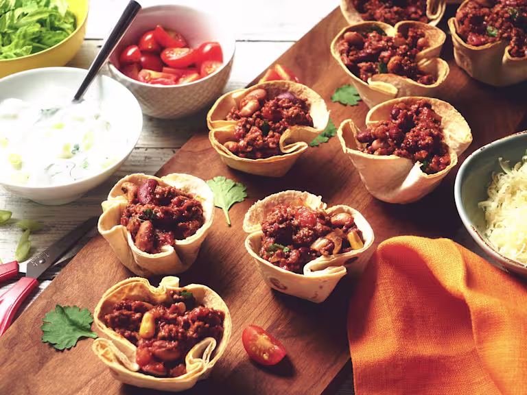 Vegetarian Quorn Mince Chilli filled in taco cups served on a wooden board with a side of cheese, yoghurt and cherry tomatoes