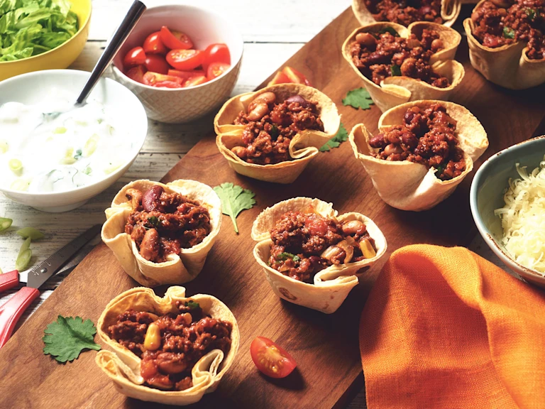 Vegetarian Quorn Mince Chilli filled in taco cups served on a wooden board with a side of cheese, yoghurt and cherry tomatoes